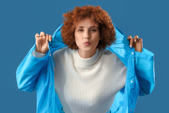Young woman in blue raincoat blowing kiss on color background