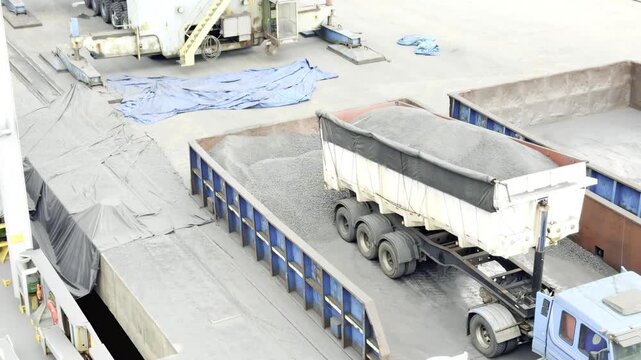 Heavy machinery in action: a commercial truck unloads black coal or gravel into a hopper as a massive crane grab lowers to transfer the industrial cargo.
