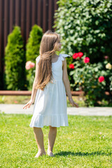 Obraz premium Cute little girl with long hair wearing a white dress stands barefoot on green grass in a garden with colorful flowers and shrubs in the background