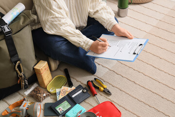 Young woman with check list of necessities for emergency backpack sitting on floor at home