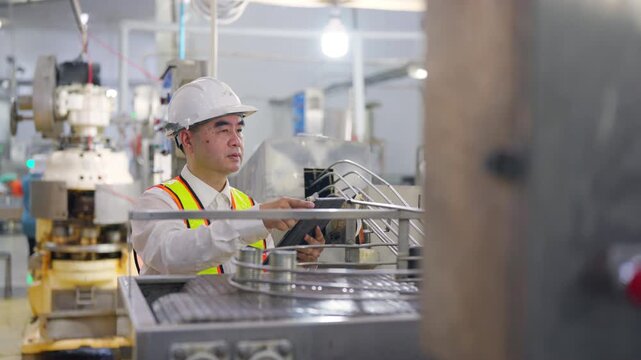 Asian man factory engineer monitoring canned food production line and inspecting manufacturing process with safety equipment inside industrial food processing plant and automated packaging facility.