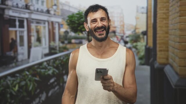 Man holding smartphone and pointing finger, laughing with bare forearm visible in a sleeveless top on a street cafe terrace; joyful interaction.