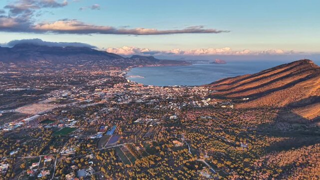 Wide aerial sunset over coastal city parallel ridge golden light bathing rooftops calm bay and distant peaks,