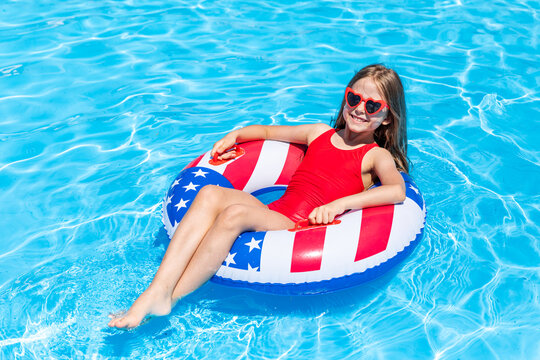 Happy smiling little girl 7-8 years old in red swimsuit and sunglasses lying on lifebuoy decorated with American flag print on blue water background. Vacation in USA concept. 4th of July pool party