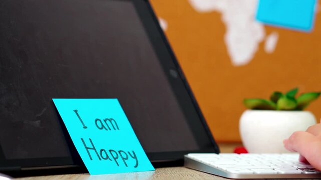 Close-up of Hand sticking blue "I am Happy" note on tablet screen while working at desk