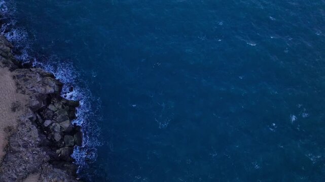 Ocean waves crashing against steep basaltic cliffs in a top-down aerial drone perspective