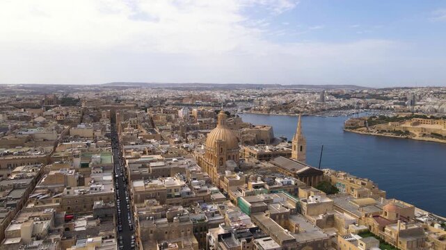 Spectacular wide aerial perspective of Valletta featuring the majestic golden dome of the Carmelite Church rising above the dense limestone cityscape, with the expansive Grand Harbour and marina beyon