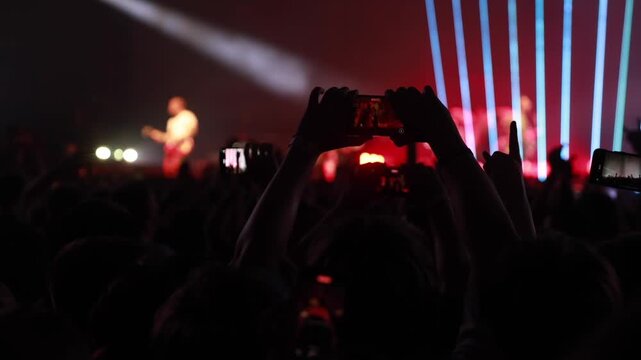 People record a musical artist on stage using smartphones during a high-energy show. Dramatic red light beams scan across the silhouetted crowd.