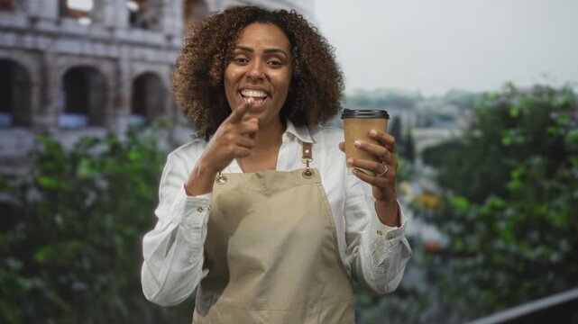 Woman baker in apron points finger to takeaway coffee cup and smiles while holding cup near coliseum; cheerful confidence.