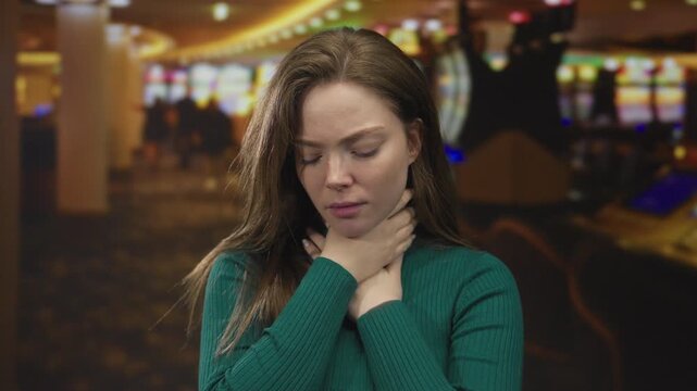 Woman clutching her throat while standing before a row of slot machines in a casino building; panic.