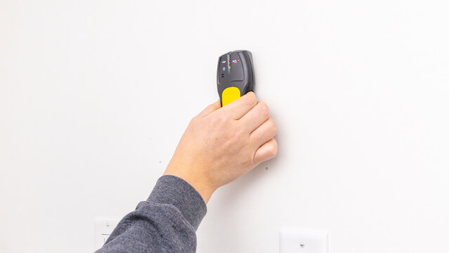 Mans hand holding yellow stud finder against white wall near electrical outlet after installation. Home improvement, checking placement, DIY verification, tool usage.