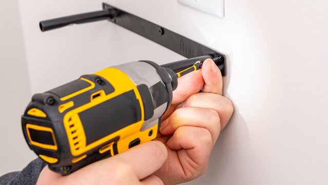 Mans hands operating yellow cordless drill attaching metal bracket to white wall with screw. Home improvement, securing fixture, DIY mounting process, tool in use.