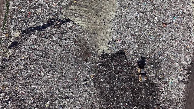 A landfill site shows large amounts of waste spread out over the ground. Heavy machinery works to manage and transport the waste. The environment has noticeable signs of garbage.