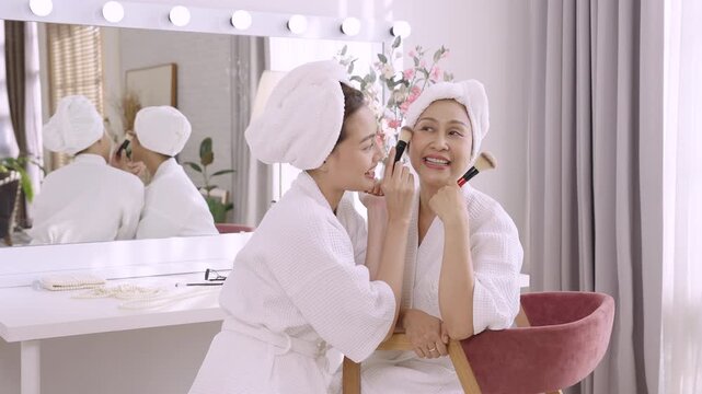 Asian daughter playfully dabbing makeup brush on smiling senior mother's cheek in slowmotion, enjoying fun skincare routine together in beauty lifestyle full of love and joy