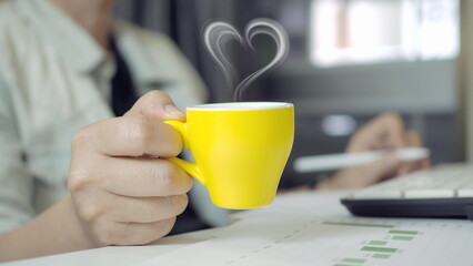 Work break lifestyle concept with yellow coffee cup and heart shaped steam. Business professional holding hot drink at desk, relaxing during work, productivity boost, morning routine.