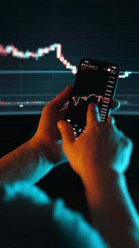 Close up of trader hands analyzing candlestick chart on smartphone with financial market graph in background. Mobile trading, investment analysis and market volatility concept in low light.