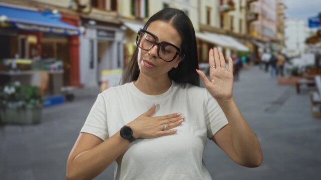 Woman wearing large glasses with hand on chest and raised hand in street; sincere pledge trust honesty.