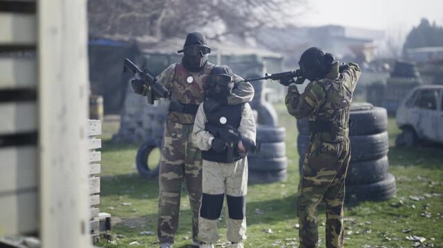 Participants in camouflage and protective gear engage in an intense air soft game on an outdoor field. They move strategically around obstacles, aiming their weapons in this action-packed scenario.