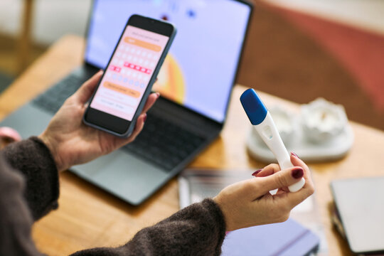 Caucasian woman holding digital pregnancy test in one hand and smartphone displaying menstrual cycle tracking app in other hand, sitting at desk with laptop in background
