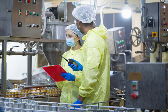 Diverse team of technicians using radio and clipboard to manage automated canning factory, Quality control inspectors in protective suits monitoring canned food production line