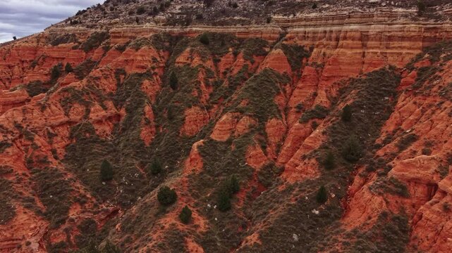 Canyon shows steep red rock formations and patches of green shrubs. The scene captures the natural shapes and textures of the land. Clouds cover parts of the sky above.