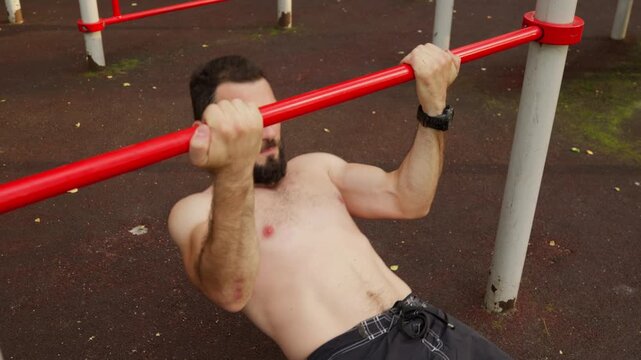 Shirtless bearded athlete performs inverted rows on a red bar at an outdoor gym. Close-up footage of a man doing calisthenics, showing strength and determination during his workout.