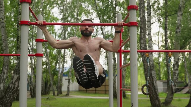 Muscular shirtless man with beard performs hanging leg raises and holds 90 degree angle on pull-up bar. Clip shows an intense ab workout at outdoor gym in park, demonstrating strength and endurance.