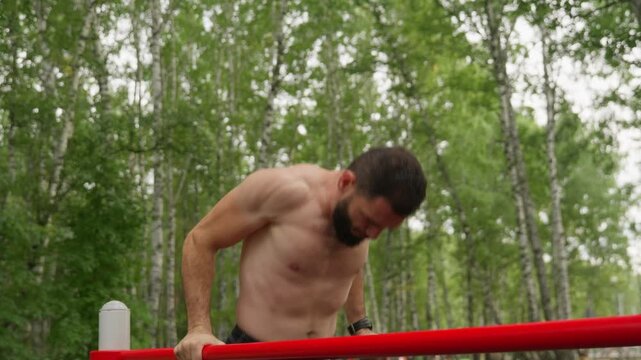A strong, shirtless man with a beard performs dips on red parallel bars. He's exercising in a green park, focusing on strength and calisthenics. Outdoor workout and fitness concept.