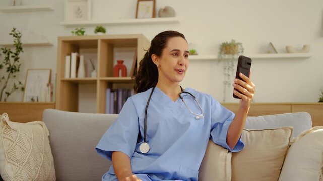 Professional female nurse in blue scrubs sitting on a couch, waving and talking during a telehealth video call on a smartphone. Friendly healthcare provider offering remote medical consultation