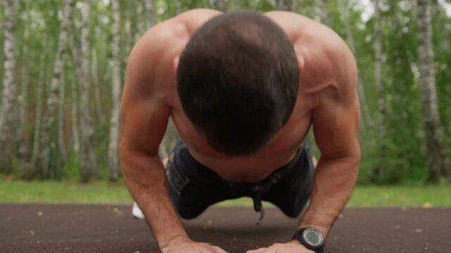 Close-up video of a shirtless, athletic man performing push-ups on a sports ground. The camera follows his movement, focusing on his muscular back and arms during an intense workout in a park.