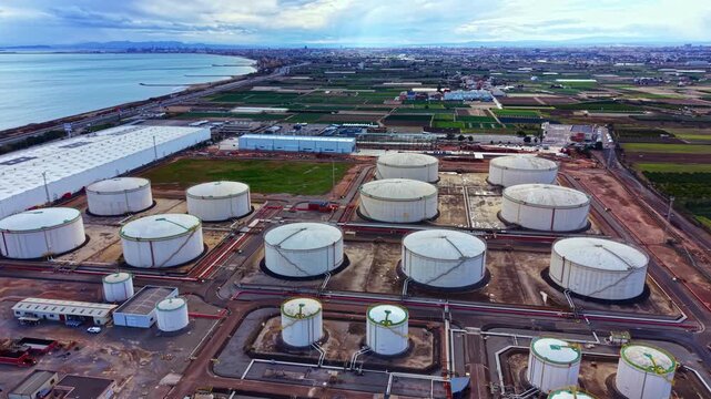Storage tanks for oil are seen from above in a coastal area. Fields and buildings are present in the background. The weather is cloudy and a blue sky is visible.