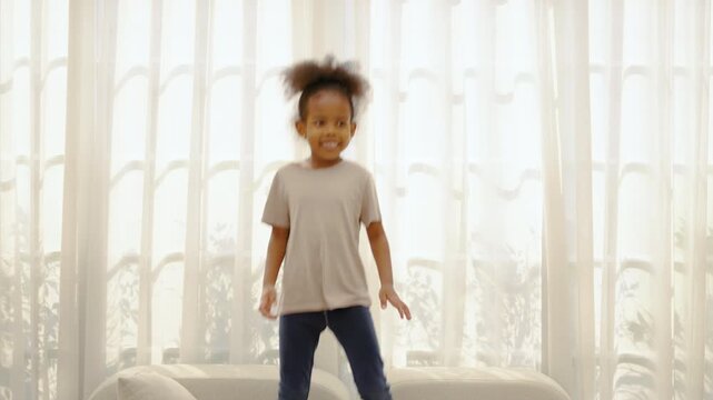 Joyful young African American girl jumping excitedly on a white sofa in a bright living room. Energetic kid playing at home during the day with sheer curtains in background. Active childhood fun.