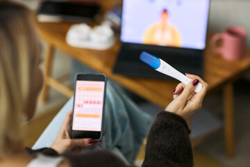 Caucasian woman holding digital ovulation test and smartphone displaying menstrual cycle tracking app, sitting indoors with laptop in background, focusing on femtech health monitoring