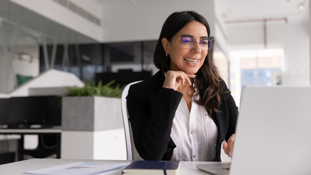 Positive middle-aged businesslady in eyeglasses working on modern laptop in office, smiling while read good news in business notification, review sales results, makes report using new, innovative tech