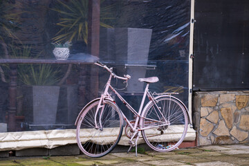 a faded pink vintage-style bicycle parked on a stone-paved walkway in front of a storefront with a dark mesh screen.