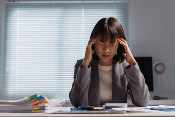 Young businesswoman feeling a severe headache while working late in the office, overwhelmed by paperwork and pressure