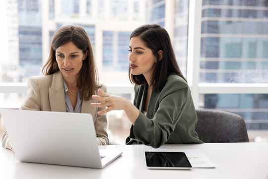 Two businesswomen work together on joint project using laptop. Woman explain or discuss key point with female colleague, collaborating on project planning, analyzing data, making decision in office