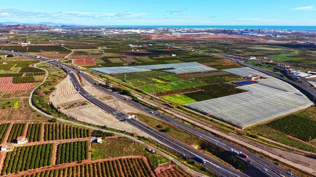 A wide landscape shows green fields, rows of crops, and highways. The scene captures farms near a coastal area with buildings in the distance under daytime sunlight.