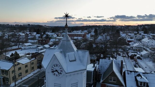 Drone view of Manheim, Pennsylvania at sunset with passing by historic white clock tower above snow-covered rooftops and small-town streets in winter. Wide shot.