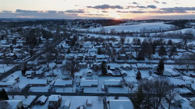 Aerial sunset over quiet American neighborhood blanketed in fresh snow, with warm golden light glowing above rooftops and rolling countryside in distance. Wide shot. PA, USA