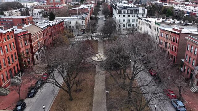 Aerial view of quiet Baltimore residential square in winter, framed by historic brick row houses and church. Leafless trees line central park, with parked cars along both sides. Backwards shot.