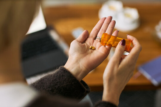 Caucasian middle aged woman holding prescription pill bottle and pouring medication into hand, sitting at desk with laptop and notebook, illustrating femtech health management