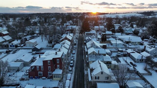 Aerial sunrise over Main Street in Manheim, Pennsylvania, with snow-covered rooftops, quiet residential homes and golden morning light glowing above the winter landscape. Birds eye flyover.