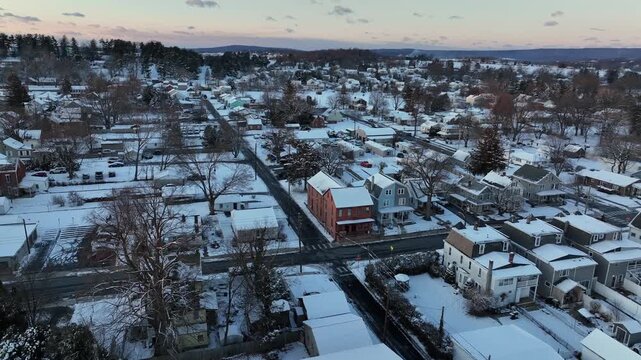 Aerial view of charming American neighborhood at sunset, with snow-covered rooftops, quiet streets and small-town homes surrounded by winter landscape and soft evening light. Wide shot. PA, USA