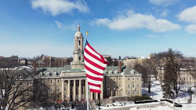 Penn State University&rsquo;s Old Main with American flag waving in foreground. Snow blankets historic campus buildings under bright blue sky in State College, Pennsylvania. Aerial view. Winter season.