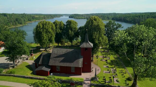 Drone shot orbits historic red wooden Sk&aring;llerud church and graveyard on scenic peninsula at N&auml;ren lakeside in Dalsland region of Sweden during sunny summer day