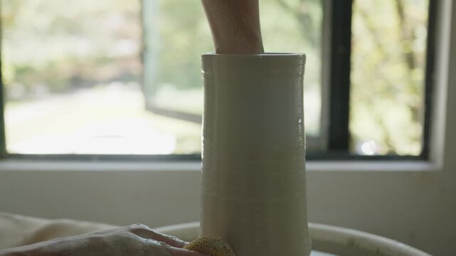 A tall vase is expertly formed on a spinning pottery wheel by an open window