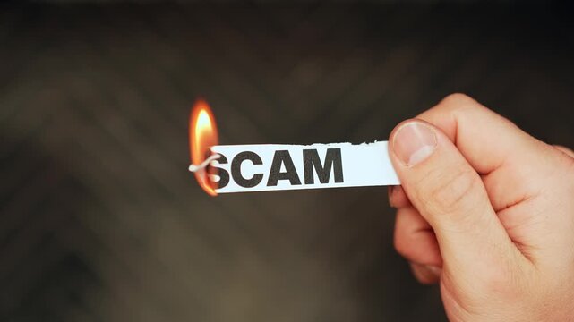 Close-up of a hand holding a paper strip labeled &ldquo;SCAM&rdquo; as the edge burns with a lighter flame on dark background, concept of fraud, deception, and risk.