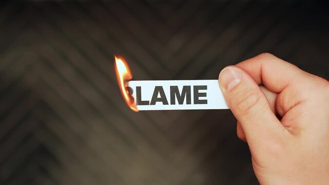 Hand holds a paper strip reading &ldquo;BLAME&rdquo; as the edge ignites from a lighter&mdash;symbolizing accountability, conflict, and letting go on a dark background with copy space.