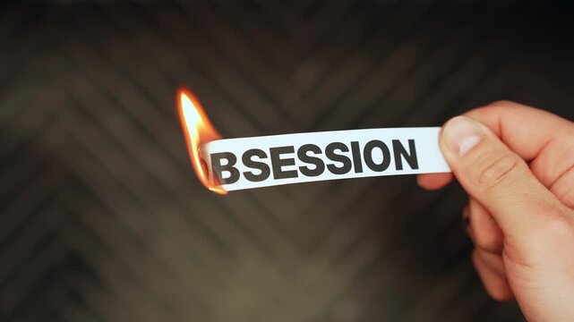 Close-up of a hand holding a paper strip labeled &ldquo;OBSESSION&rdquo; as the edge ignites from a lighter&mdash;symbolic letting go, habit change, and mindset shift on a dark background.